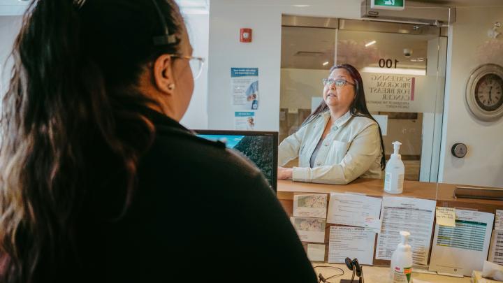 dark-haired women in a gray jacket speaking to a dark haired women with a headset on as she hands her a pink scrub shirt in a clinic office reception.